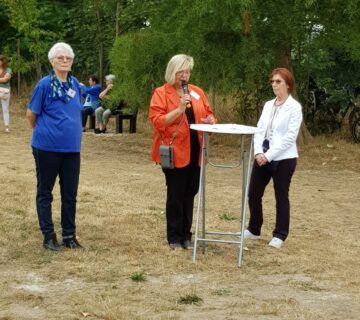 Drei Frauen stehen auf einer Wiese. Die Frau in der Mitte steht an einem Tisch und hält ein Mikrofon in der Hand.