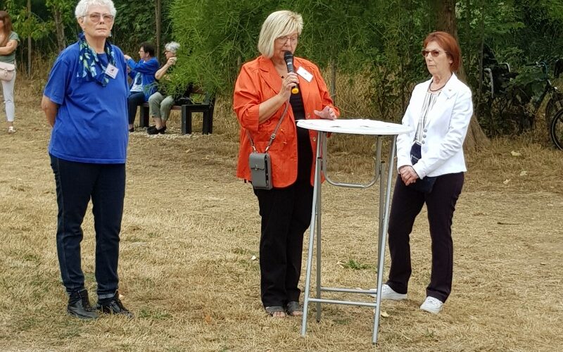 Drei Frauen stehen auf einer Wiese. Die Frau in der Mitte steht an einem Tisch und hält ein Mikrofon in der Hand.