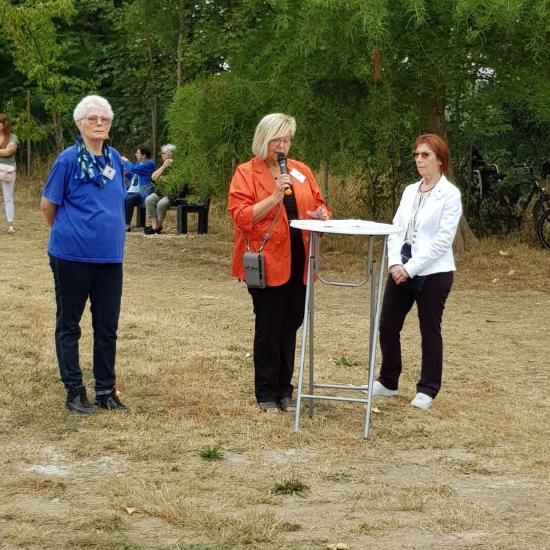 Drei Frauen stehen auf einer Wiese. Die Frau in der Mitte steht an einem Tisch und hält ein Mikrofon in der Hand.
