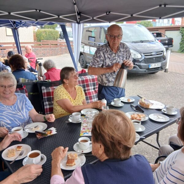 Mehrere Frauen sitzen am Tisch und trinken Kaffee und essen Kuchen. Ein Mann steht am Tisch.